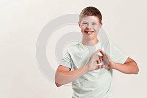 Joyful disabled boy with Down syndrome smiling at camera, making heart shape with his hands while standing isolated over