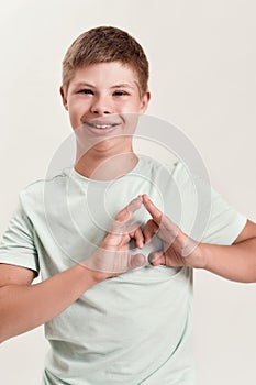 Joyful disabled boy with Down syndrome smiling at camera, making heart shape with his hands while standing isolated over