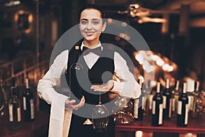 Joyful elegant waitress holding bottle of red wine and glasses, standing near bar.