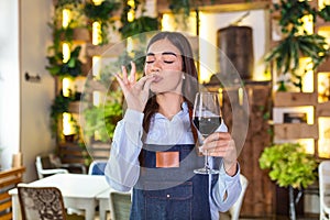 Joyful elegant waitress holding glass of red wine showing delicious sign , standing in restaurant. Sommelier tasting wine in