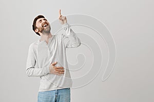 Joyful emotive young european male bending backwards while laughing out loud and holding belly, pointing up with index