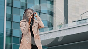 Joyful energetic young woman dancing outdoors listening to music with headphones singing using smartphone as microphone