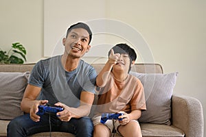 Joyful and excited Asian dad and son with joysticks are playing video games at home together