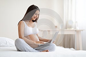 Joyful expecting woman relaxing in bed with laptop