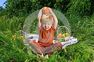 nice and funny fair-haired kid holds an orange over his head, basket with fruits on green grass