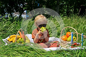 funny fair-haired toddler with sunglasses holds an apple, basket with fruits on green grass