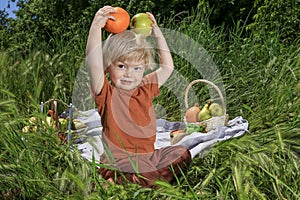 cute fair-haired toddler holds an apple and orange, basket with fruits on green grass