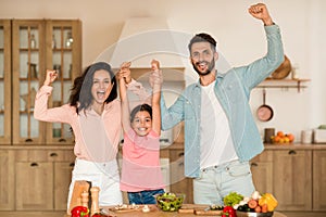 Joyful family holding hands and shaking fists, gesturing yes while preparing dinner together in kitchen