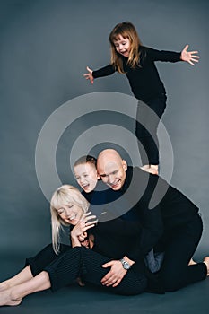 Joyful family mother, father and two little children in black clothes having fun on floor with dark background