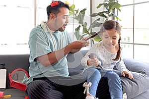 Joyful father playing with his little adorable daughter together, girl doing makeup to her dad by cosmetic, sitting on sofa in