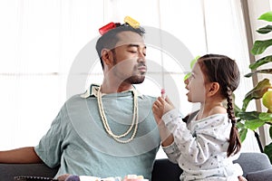 Joyful father playing with his little cute daughter together, girl doing makeup to her dad by cosmetic during sitting on sofa in