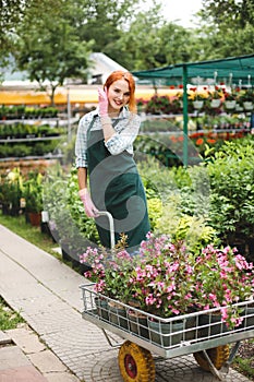 Joyful gardener girl in apron and pink gloves looking in camera working with flowers in cart in garden at sprin time