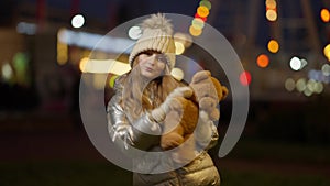 Joyful girl dancing with teddy bear in slowmo with colorful Christmas lights flashing at background on city street