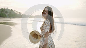 Joyful girl in flowery dress posing, flirting on camera at tropical beach in sunrise light. Young pretty woman in straw