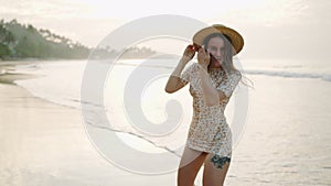 Joyful girl in flowery dress running, posing at tropical beach in sunrise light. Young pretty woman in straw hat has fun