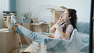 Joyful girl homeowner talking on mobile phone after relocation relaxing on packed furniture