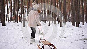 Joyful girl running pulling sledge on white snow in winter park. Back view wide shot happy excited Caucasian teenager