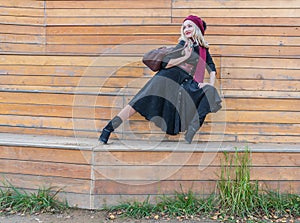 A joyful girl sits on a summer theater bench, made of wood in a burgundy coat and biret, an adult looking at the camera