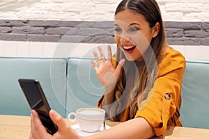 Joyful girl video calling friend on smart phone she greets with her hand. Young woman using phone camera for a chat with her