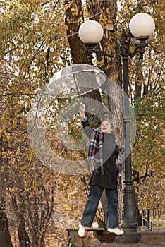 Joyful girl with wool scarf holds umbrella over her on outstretched arm. Woman stands under lantern in fall park