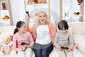 Joyful grandmother with cheerful grandchildren playing on game console sitting on couch.