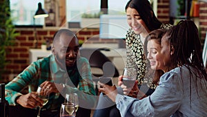 Joyful guests in living room watching internet clips on mobile phone while drinking wine