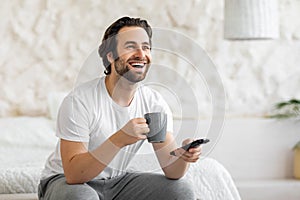 Joyful guy sitting on bed, watching TV and drinking coffee