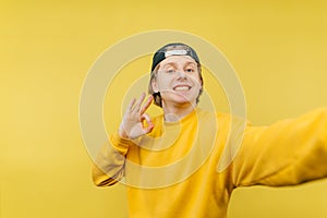 Joyful guy in a yellow sweatshirt and cap takes a selfie with a smile on his face and shows an OK gesture, isolated on a yellow