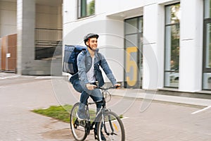 Joyful handsome young delivery man with thermo backpack riding bicycle in city street on blurred background of office