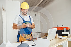 Joyful handyman in workwear and hardhat smiling while using smartphone, standing indoors during renovation work