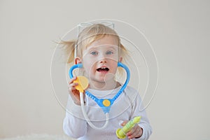 Joyful happy baby girl imagines as doctor at hospital using toy stethoscope and syringe.