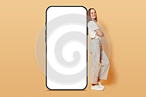 Joyful happy delighted woman leaning on big samrtphone with empty screen standing with crossed arms isolated on beige background