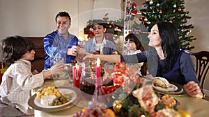 Joyful happy family clinking juice glasses talking dining in living room on Christmas. Positive Middle Eastern man boys