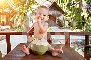 A joyful and happy little one year old child sits on a wooden table and drinks coconut milk from fresh green coconut through a
