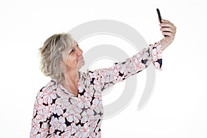 Joyful happy senior elderly blonde woman in flowers blouse takes picture selfie with cell smartphone on white background