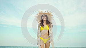 Joyful happy travelr woman in fashionable swimwear and straw hat goes on camera wiht sea view on background.