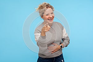 Joyful happy woman with short curly hair in sweatshirt pointing at camera, holding her stomach and laughing at crazy joke