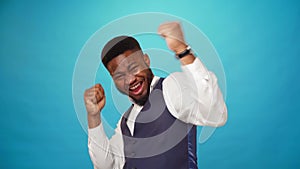 Joyful happy young african american guy celebrates success, he is winner, blue background