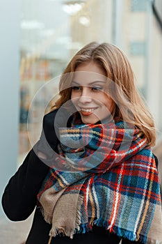 Joyful happy young woman in a fashionable warm wool scarf in a stylish black coat in black gloves is standing and smiling