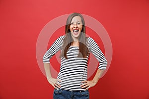 Joyful happy young woman in striped clothes keeping mouth wide open, standing with arms akimbo isolated on bright red