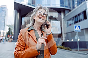 Joyful hoary haired senior lady talks on cellphone on modern city street