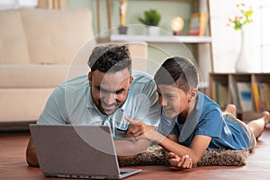 Joyful indian father with son watching laptop by lying on floor at home - concept of cyberspace, Joyful Bonding and
