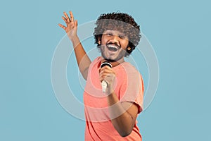 Joyful indian guy singing karaoke on blue, holding microphone