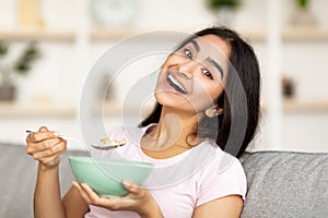 Joyful Indian lady eating granola or cereal with milk on comfy couch at home, smiling at camera