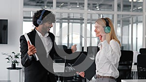 Joyful individuals wearing wireless headphones dancing and having fun in office.