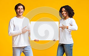 Joyful interracial couple pointing at white blank placard in their hands