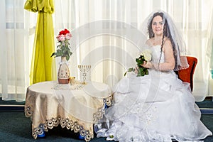 A joyful Jewish bride with her face covered with a veil with a bouquet of white roses sits in a synagogue before