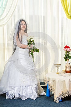 A joyful Jewish bride with her face covered with a veil and a bouquet of white roses is standing in the synagogue before