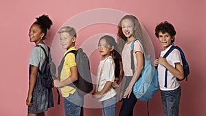 Joyful kids with backpacks going to school one after another on pink background