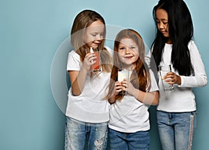Joyful kids girls friends in blue jeans and white t-shirts stand with glasses of water, milk and juice in hands
